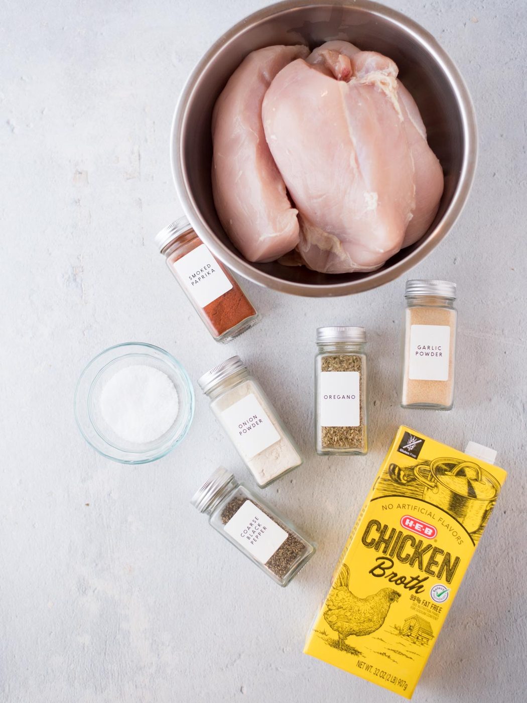Raw chicken breasts in a metal bowl, a carton of chicken broth, small jars of spices, and a bowl of salt are arranged on a light surface—everything you need to make flavorful instant pot shredded chicken breast.