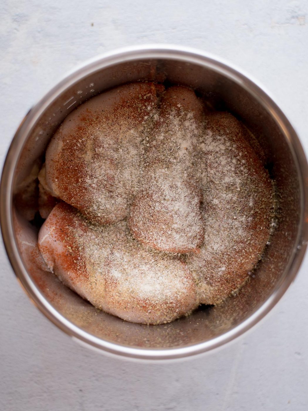 Raw chicken breasts in a metal bowl, sprinkled with various seasonings including salt, pepper, and paprika, on a light-colored surface—evenly prepared for making instant pot shredded chicken breast.