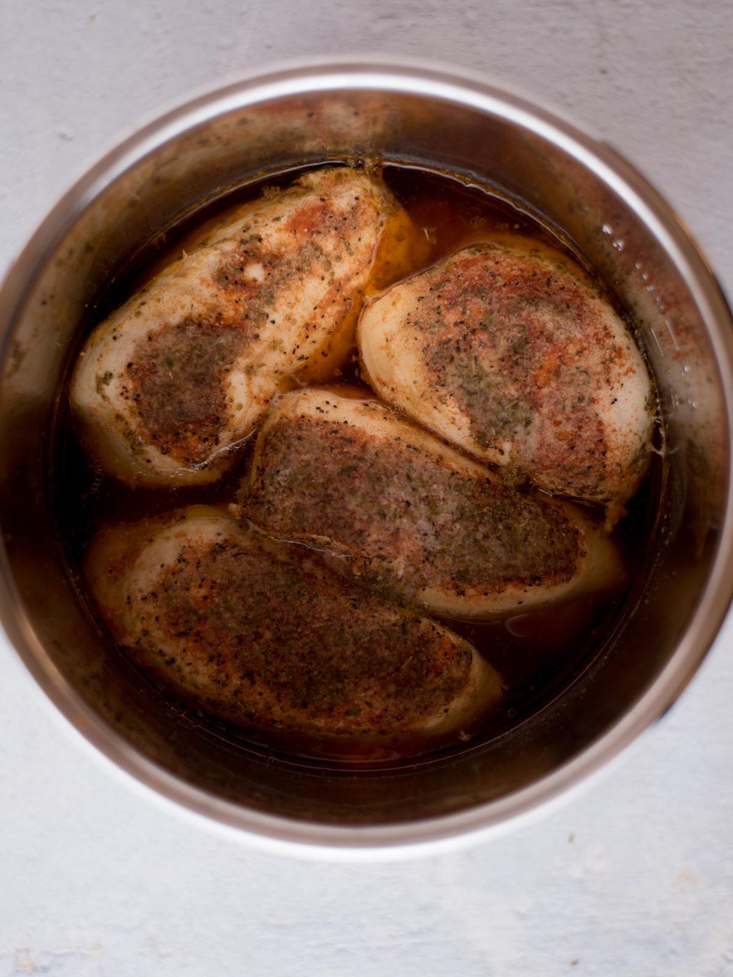 Four seasoned chicken breasts sit in broth inside a stainless steel pot, viewed from above. Well-balanced for instant pot shredded chicken breast, the chicken is cooked and covered with herbs and spices, making it straightforward to prepare shredded chicken in the instant pot. The pot is placed on a light surface.