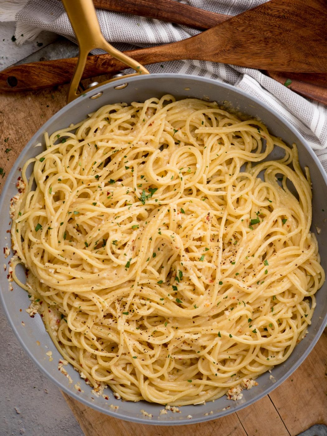 A skillet filled with freshly cooked spaghetti, lightly coated in a creamy sauce with a sprinkle of herbs and black pepper on top. A wooden spatula and a striped cloth are visible in the background.
