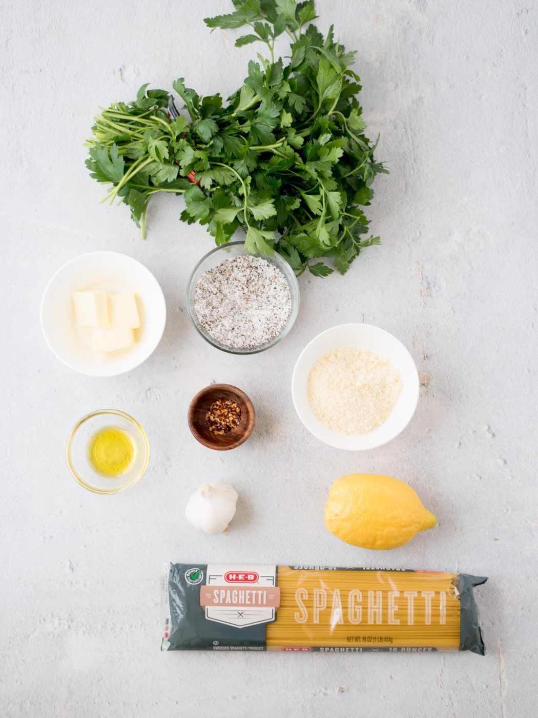 A bundle of spaghetti, fresh parsley, lemon, garlic bulb, a small bowl of parmesan, a small bowl of butter cubes, a small wooden bowl of red pepper flakes, and a small glass bowl of olive oil are arranged on a light surface.