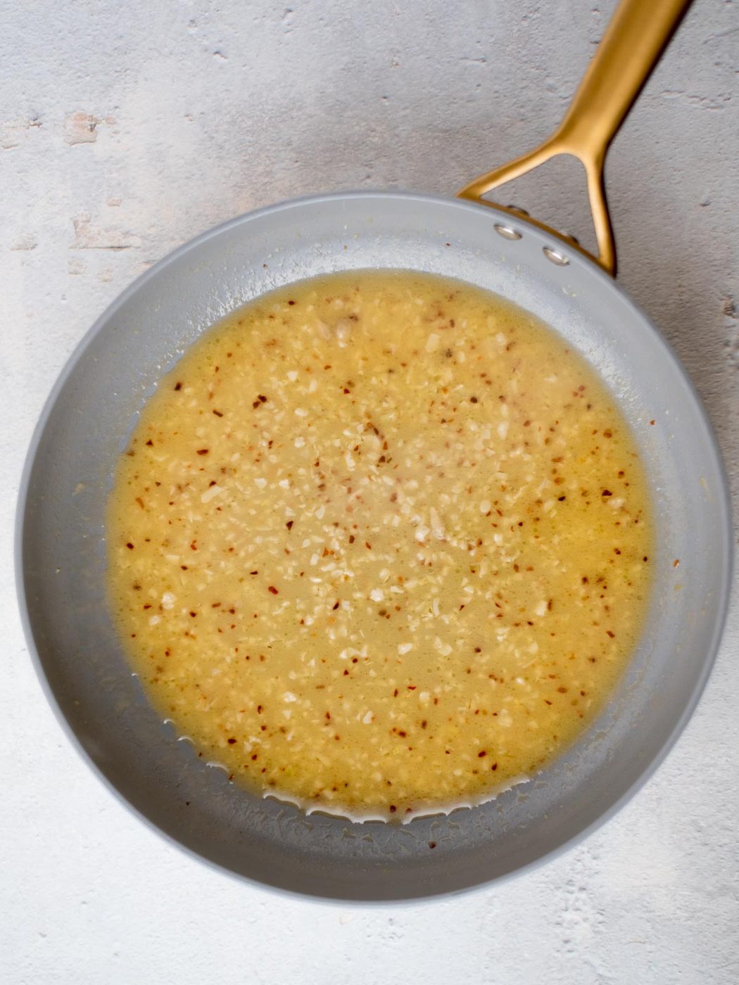 A frying pan with a gray interior and a gold handle contains a creamy, speckled sauce. The sauce appears to be a butter, garlic, and mustard mixture, with visible small pieces of garlic or seasonings dispersed throughout. The background is light gray.