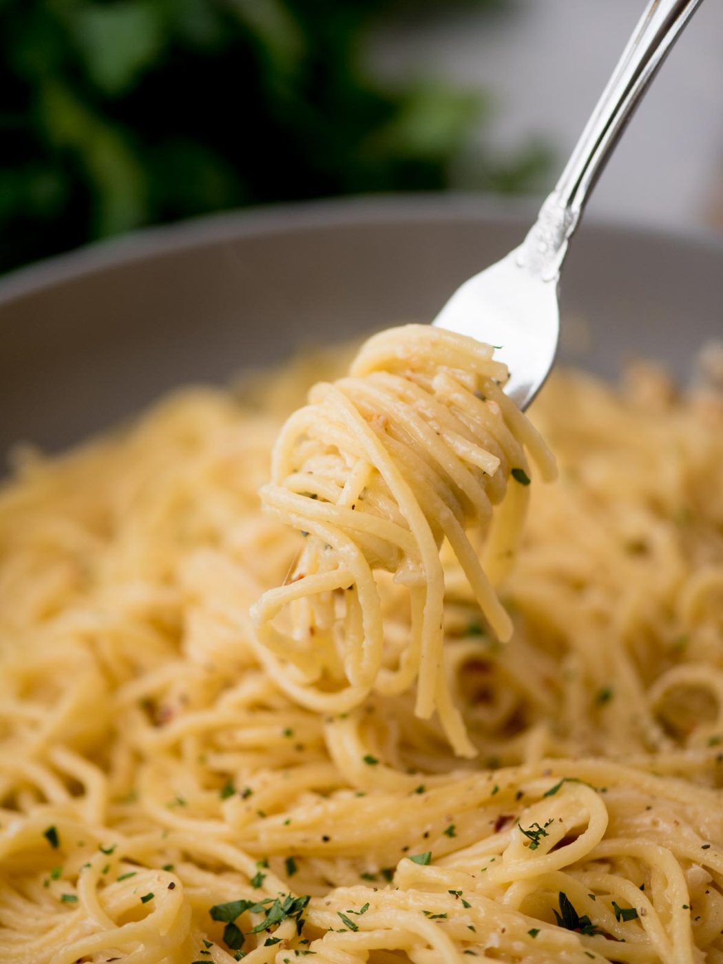 A fork twirls a portion of creamy spaghetti over a bowl. The pasta is topped with finely chopped herbs, giving it a fresh, appetizing look. The background is slightly blurred, focusing on the pastas texture.