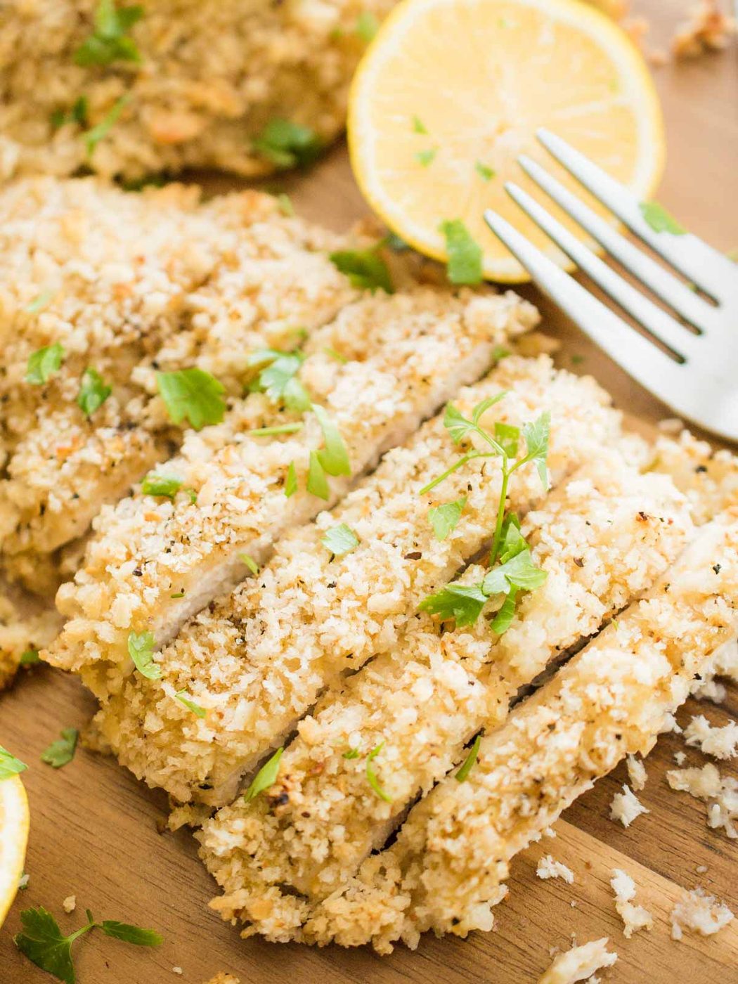 up close shot of sliced lemon pepper chicken on a cutting board with a fork, lemons, and parlsey
