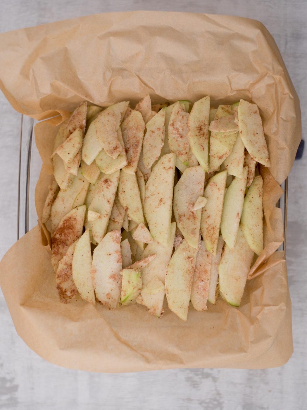 Sliced apples seasoned with cinnamon on parchment paper in a baking dish.