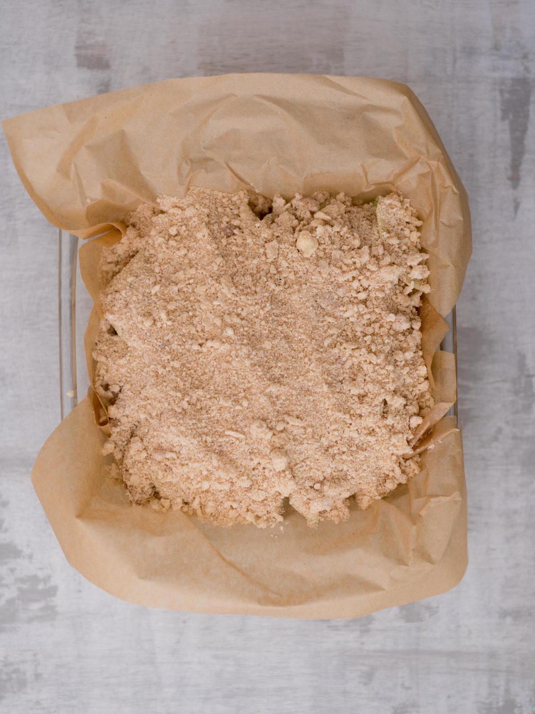 A baking dish lined with brown parchment paper is filled with an unbaked, crumbly, cinnamon-colored topping, ready to be baked. The background is a light gray surface.