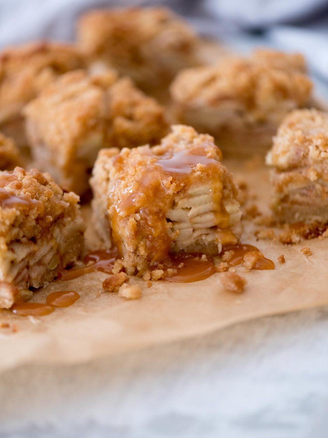 Close-up of apple crumble bars with layers of apple slices and a crumbly topping. Drizzled with caramel sauce, the bars sit on a piece of parchment paper, creating a satisfying and inviting dessert scene.