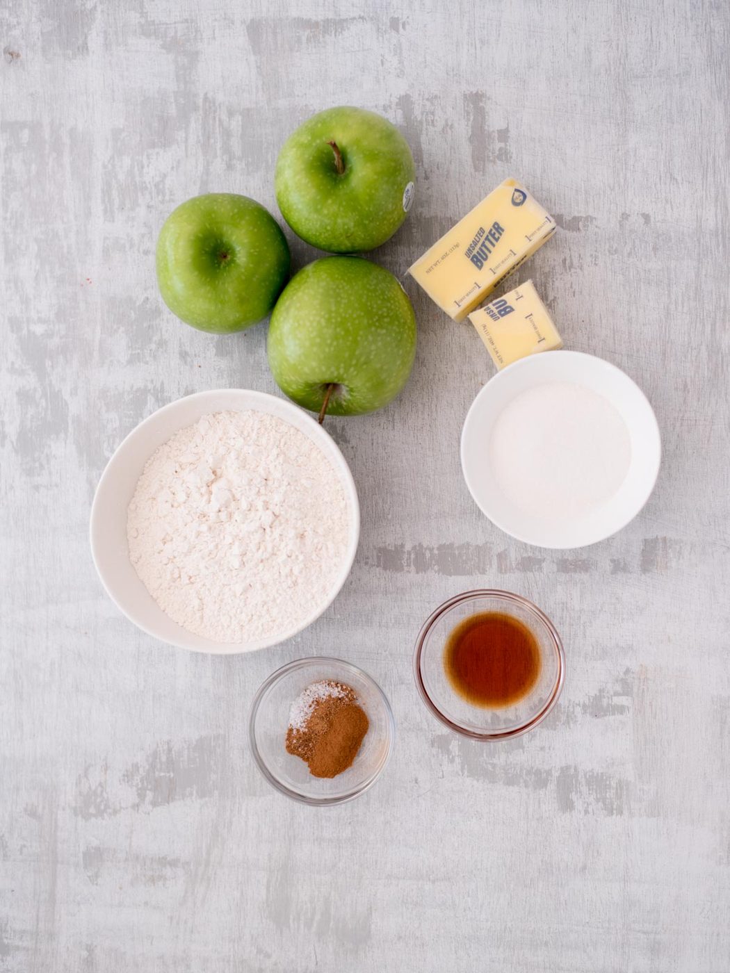 Top-down view of baking ingredients on a gray surface, including three green apples, two sticks of butter, a bowl of flour, a bowl of sugar, and small bowls with spices and vanilla extract.