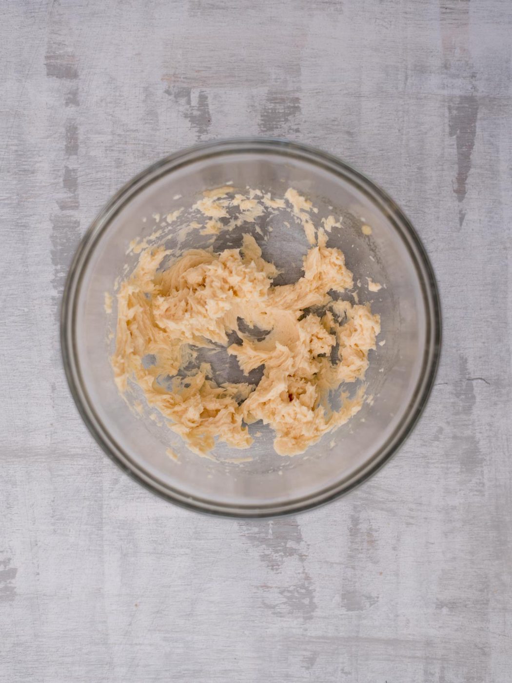 A glass bowl containing creamy, partially mixed batter is placed on a light gray wooden surface. The batter is unevenly blended, with some visible lumps.