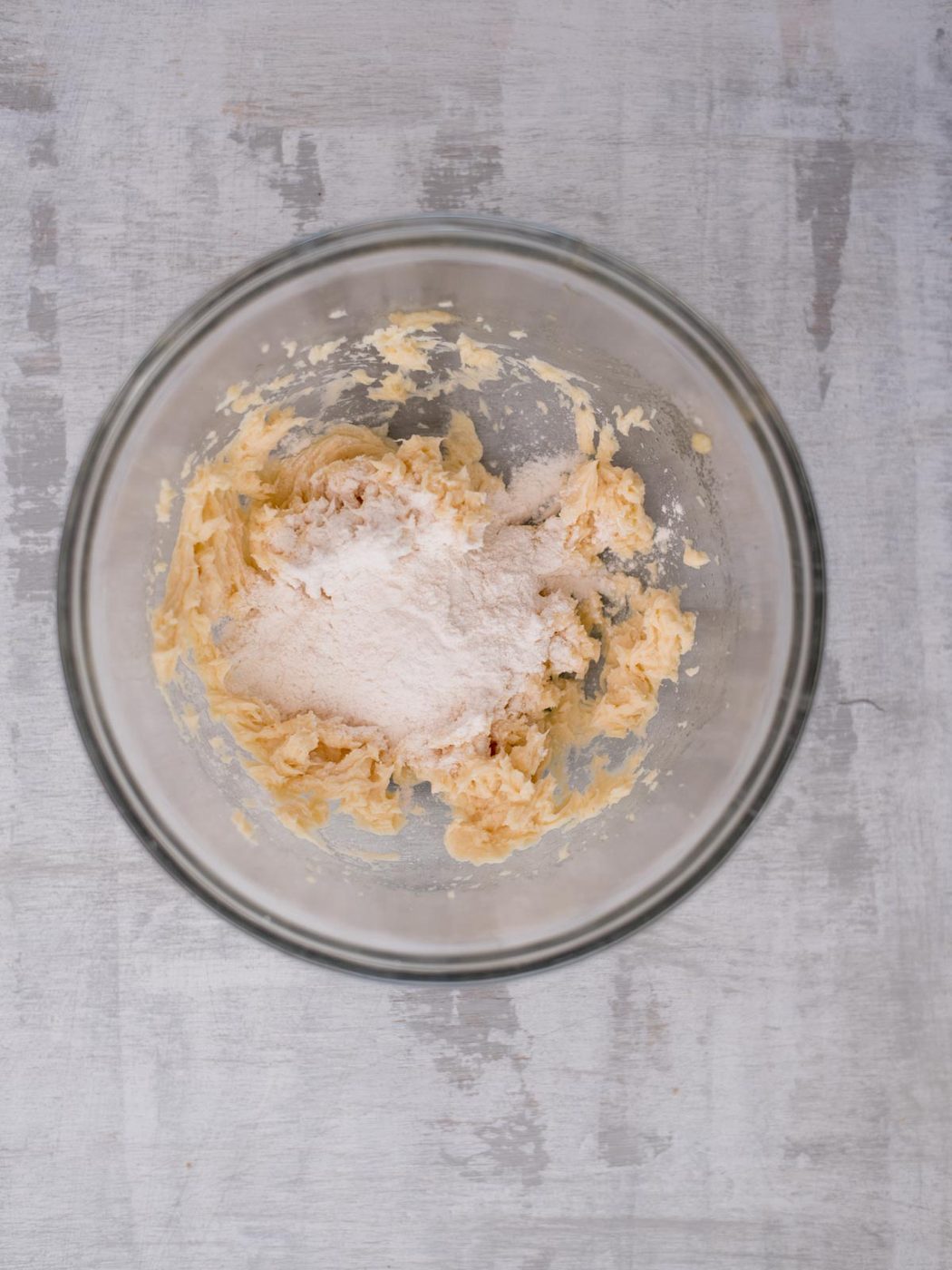 A glass bowl contains partially mixed dough with flour and softened butter on a textured gray surface. The ingredients are in the early stages of forming a cohesive dough mixture.