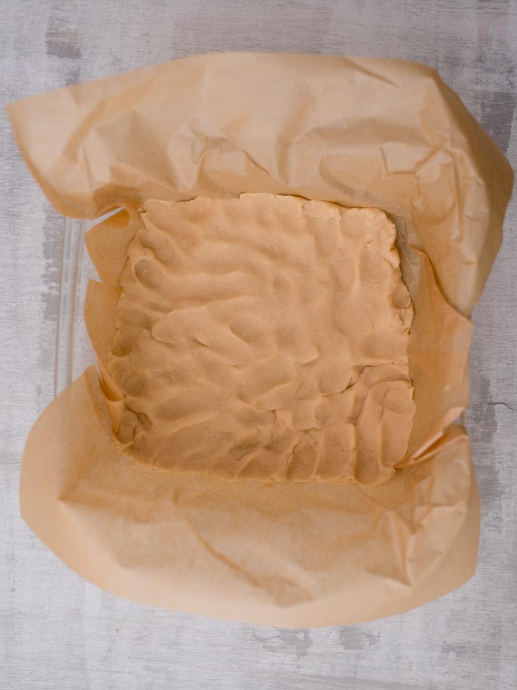 A square baking pan lined with parchment paper containing an evenly pressed layer of raw shortbread dough. The dough has a slightly textured surface. The pan is placed on a gray countertop.