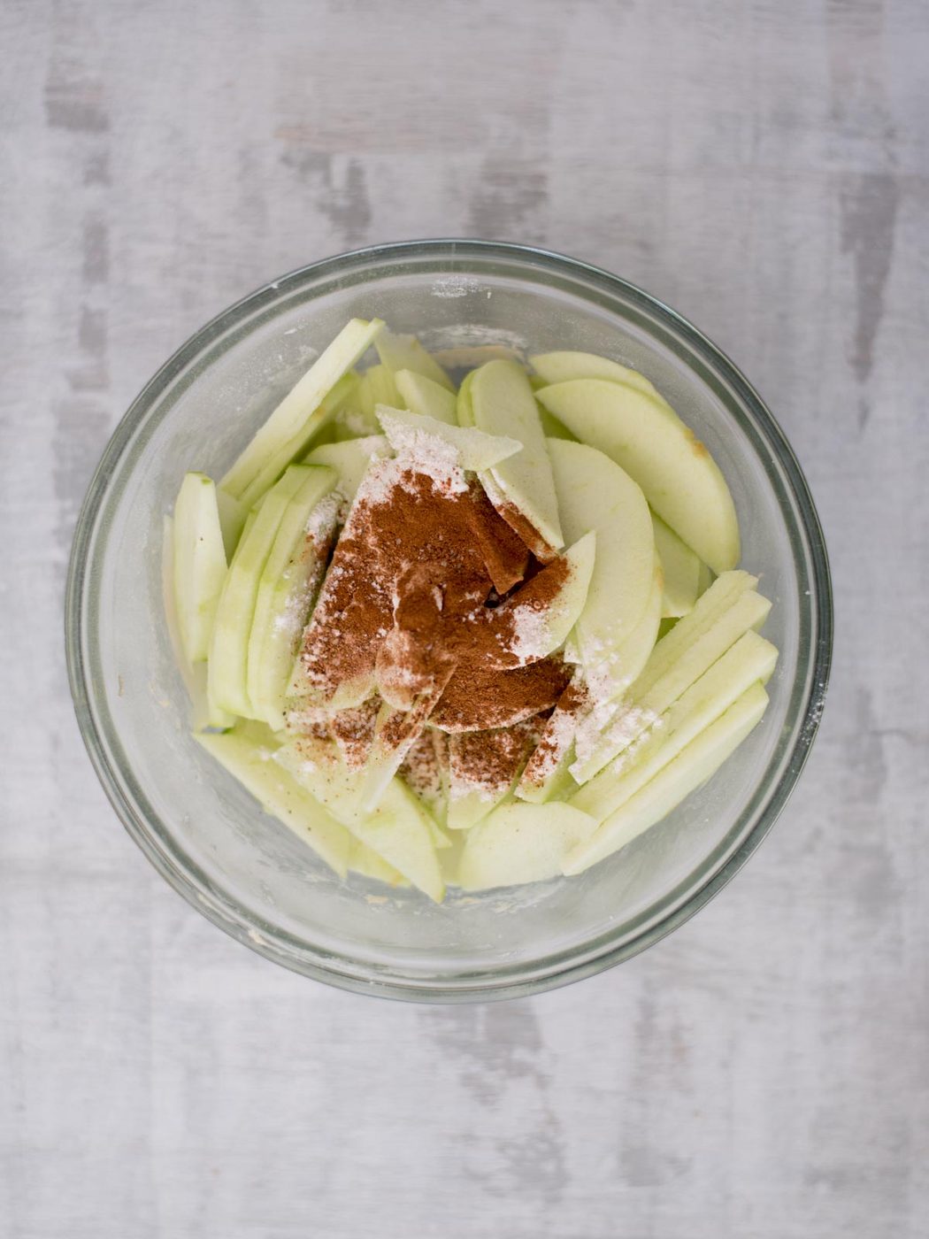 A glass bowl containing sliced green apples, sprinkled with cinnamon and flour, placed on a light gray surface.