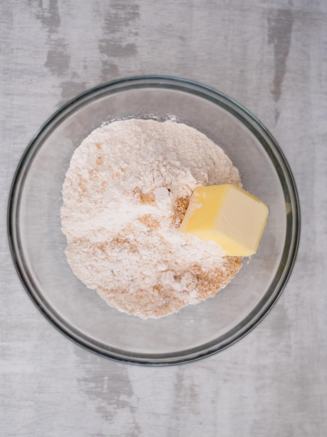 A glass bowl contains baking flour and a block of butter, ready for mixing, placed on a gray surface.