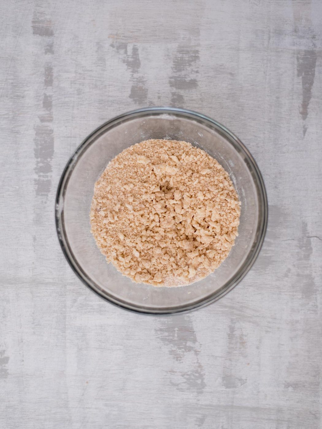 A glass bowl filled with a crumbly mixture of light brown, dry ingredients sits on a textured gray surface, viewed from above.