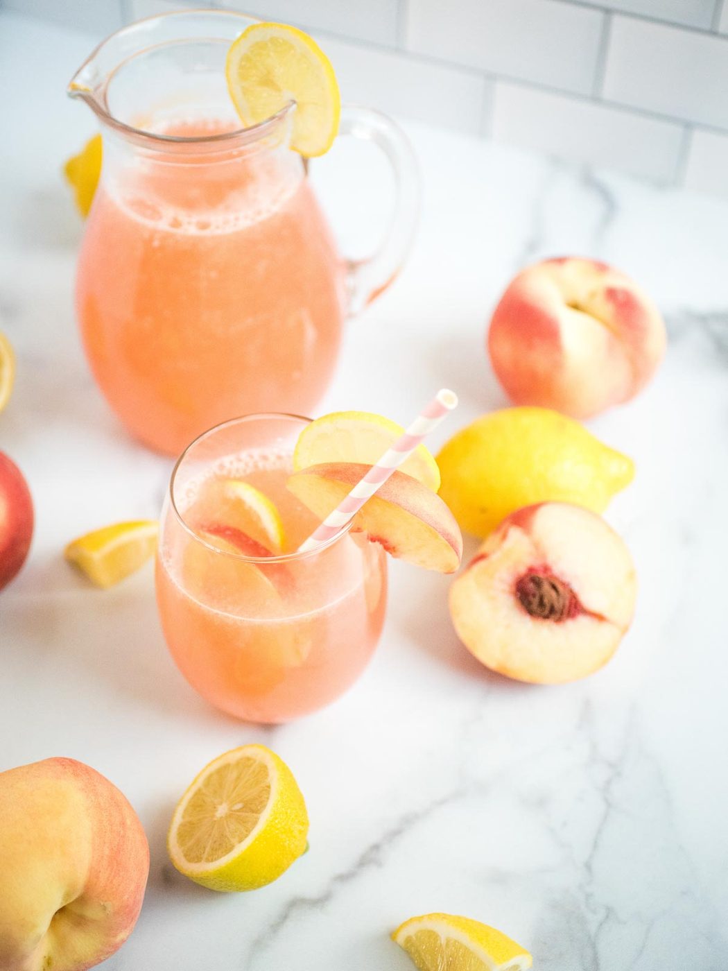 a glass of peach lemonade surrounded by lemons and peaches with the large pitcher of peach lemonade in the background