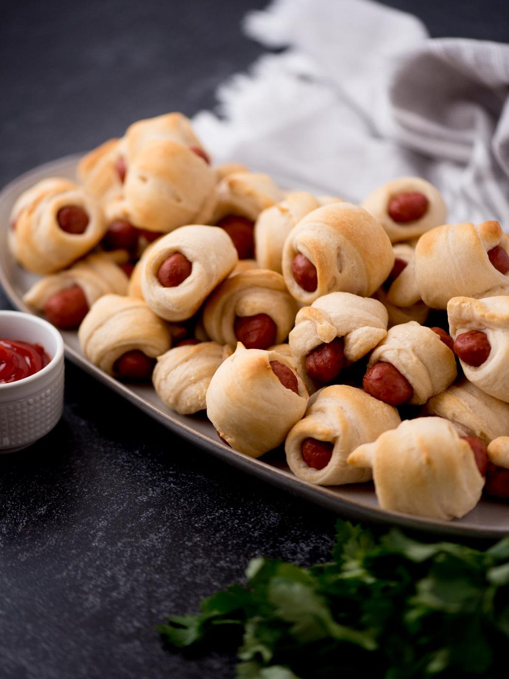 Lil smokies pigs in a blanket stacked on top of each other on a platter with a small bowl of ketchup.