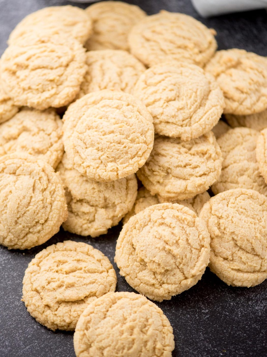 A pile of golden, round, soft vanilla pudding cookies is arranged on a dark surface. The cookies have a slightly cracked texture on top and look freshly baked.