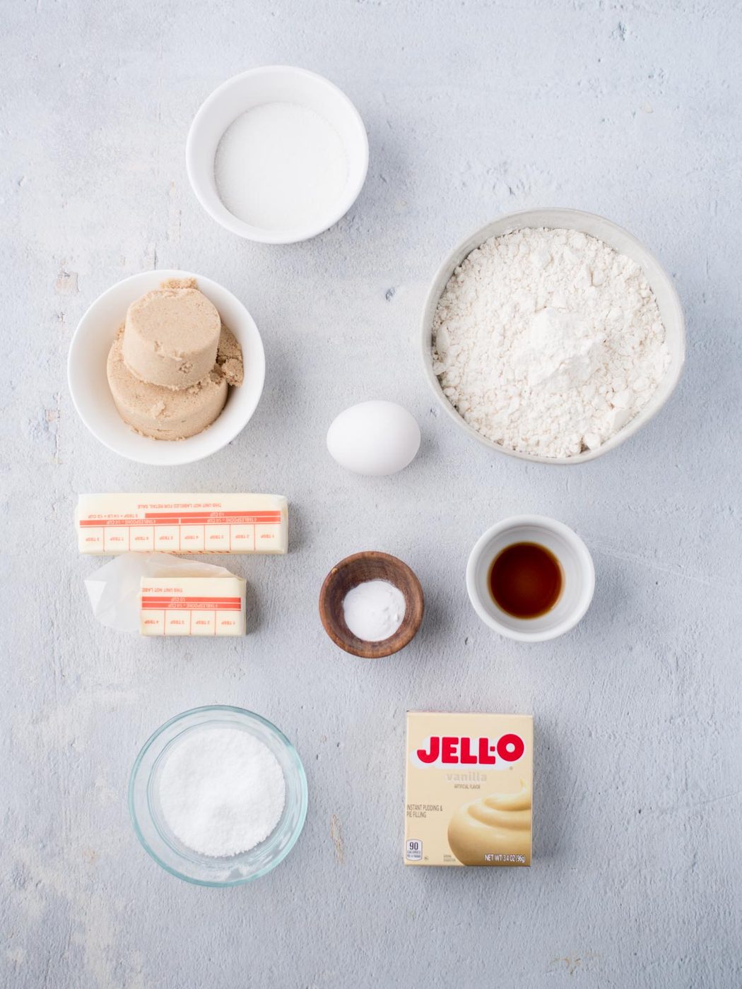 A top-down view of baking ingredients neatly arranged on a light surface, ready to make satisfying vanilla pudding cookies, featuring flour, egg, sugar, brown sugar, butter, baking powder, baking soda, vanilla extract, and JELL-O pudding mix.