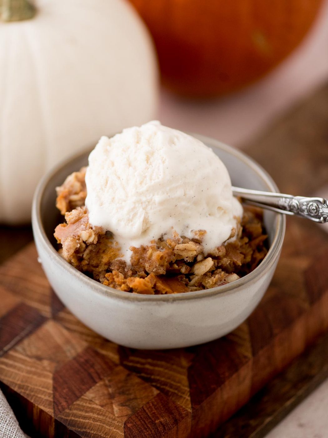 A bowl of pumpkin crisp topped with a scoop of vanilla ice cream on a wooden board. In the background, a white pumpkin is partially visible. A spoon rests in the bowl.