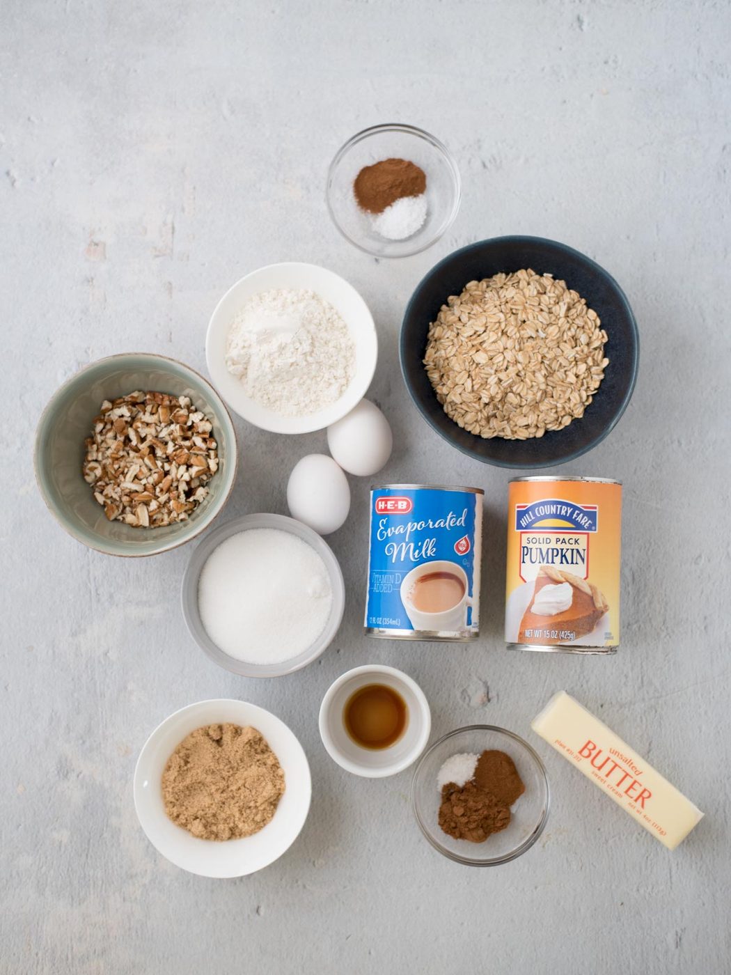Various baking ingredients arranged on a light surface: rolled oats, chopped nuts, flour, sugar, evaporated milk, pumpkin puree, eggs, butter, brown sugar, vanilla extract, and spices in small bowls.