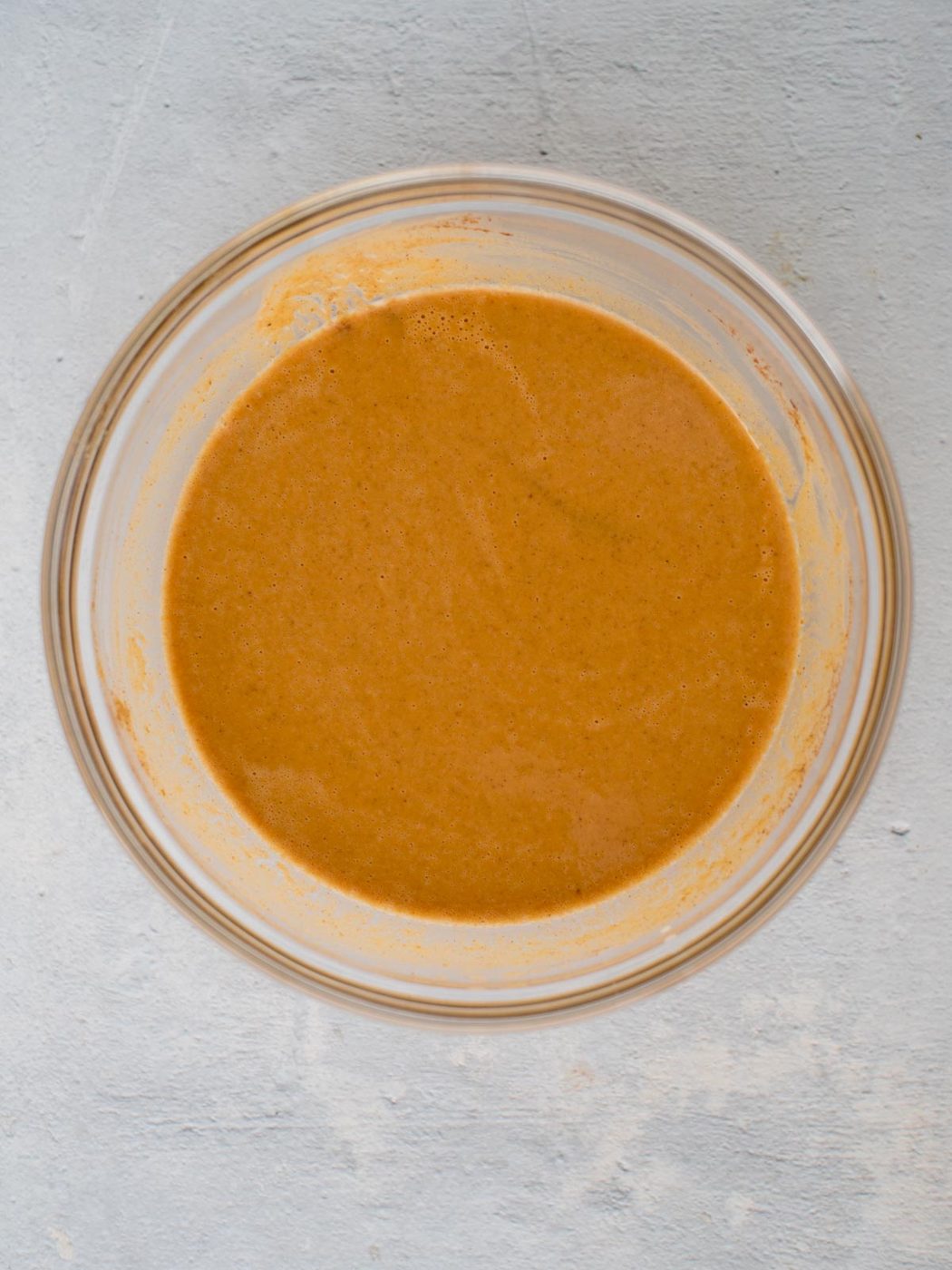 A glass bowl filled with smooth, creamy light brown batter or sauce, placed on a light gray textured surface.