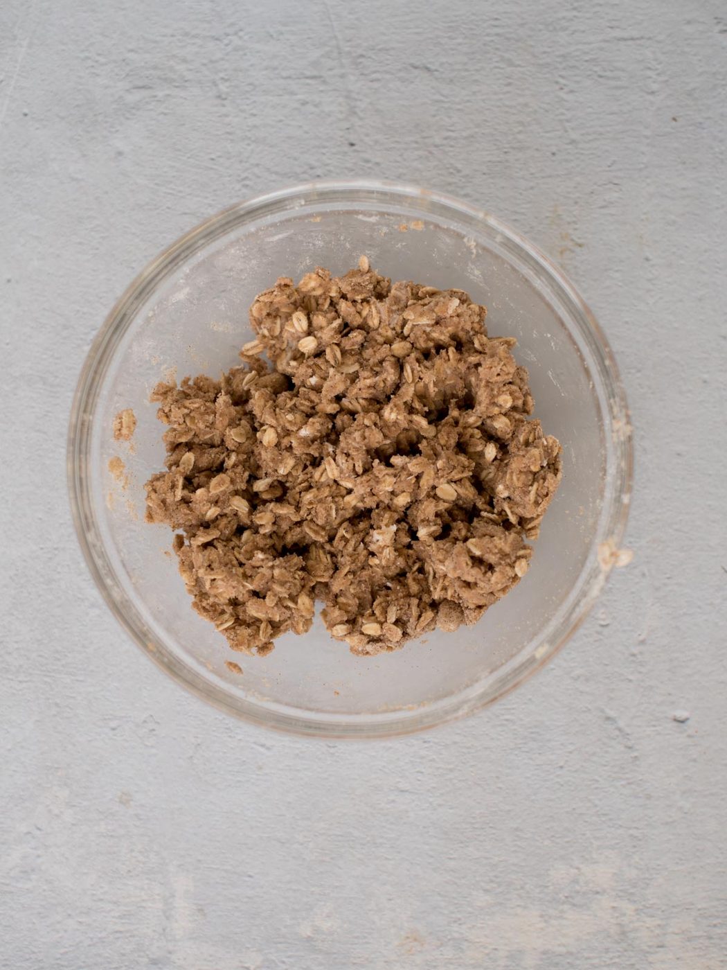 A clear glass bowl filled with crumbly brown cookie dough, likely containing oats, placed on a light gray textured surface.