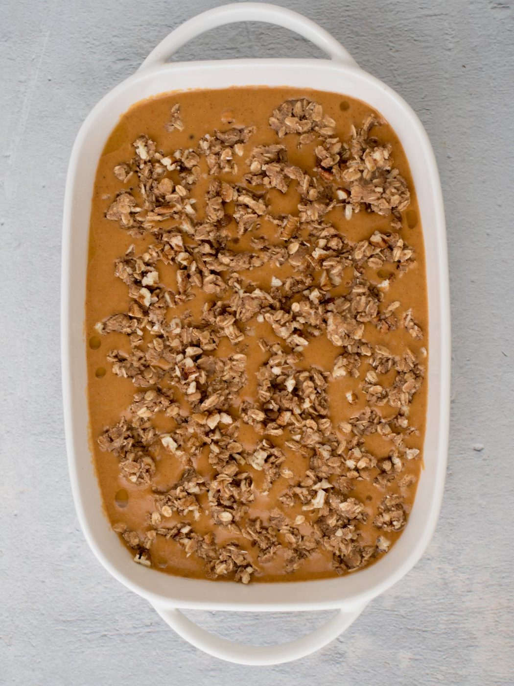 A white baking dish filled with a caramel-colored dessert topped with a crumbly nut mixture, possibly pecans, against a light gray background.