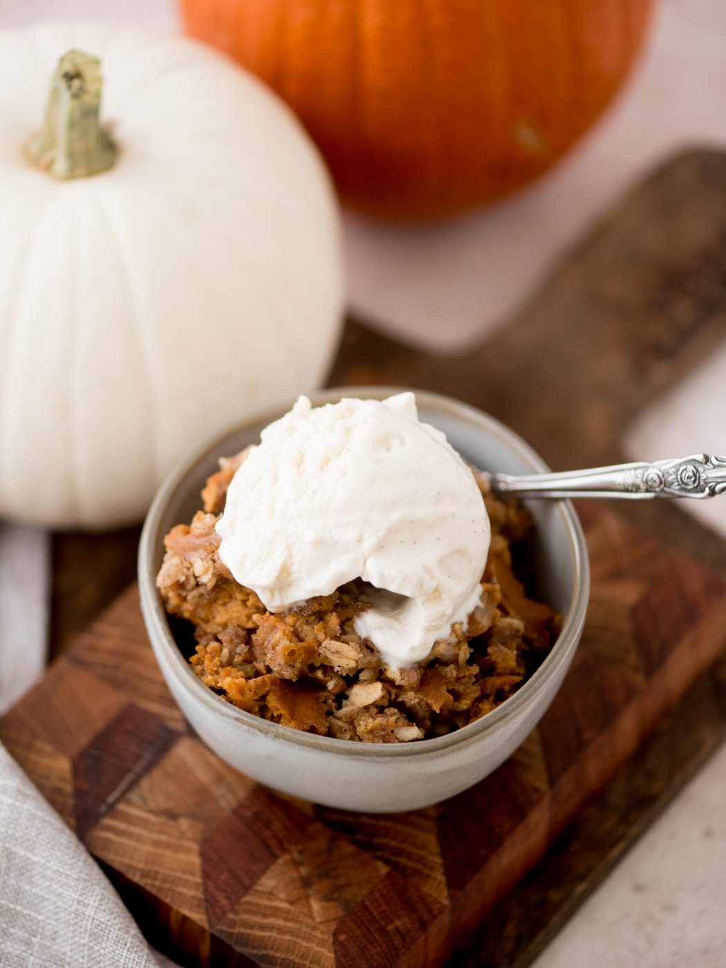 A bowl of pumpkin crisp topped with a scoop of vanilla ice cream sits on a wooden board. Two pumpkins, one white and one orange, are in the background. A silver spoon is resting on the bowls rim.