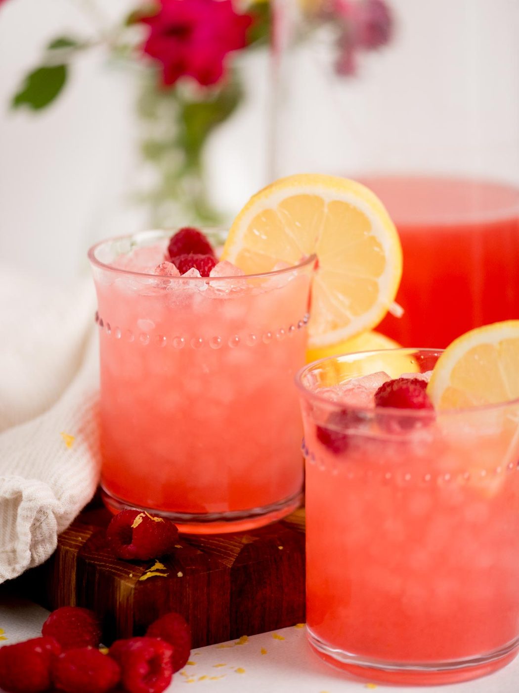 Two glasses of pink raspberry lemonade with ice, garnished with lemon slices and raspberries, sit on a wooden board. Fresh raspberries and a white cloth are nearby, with blurred flowers in the background.