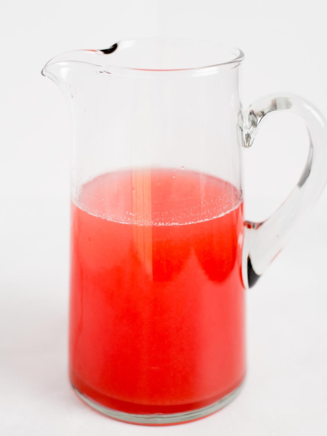 A clear glass pitcher filled halfway with bright red liquid, set against a plain white background.