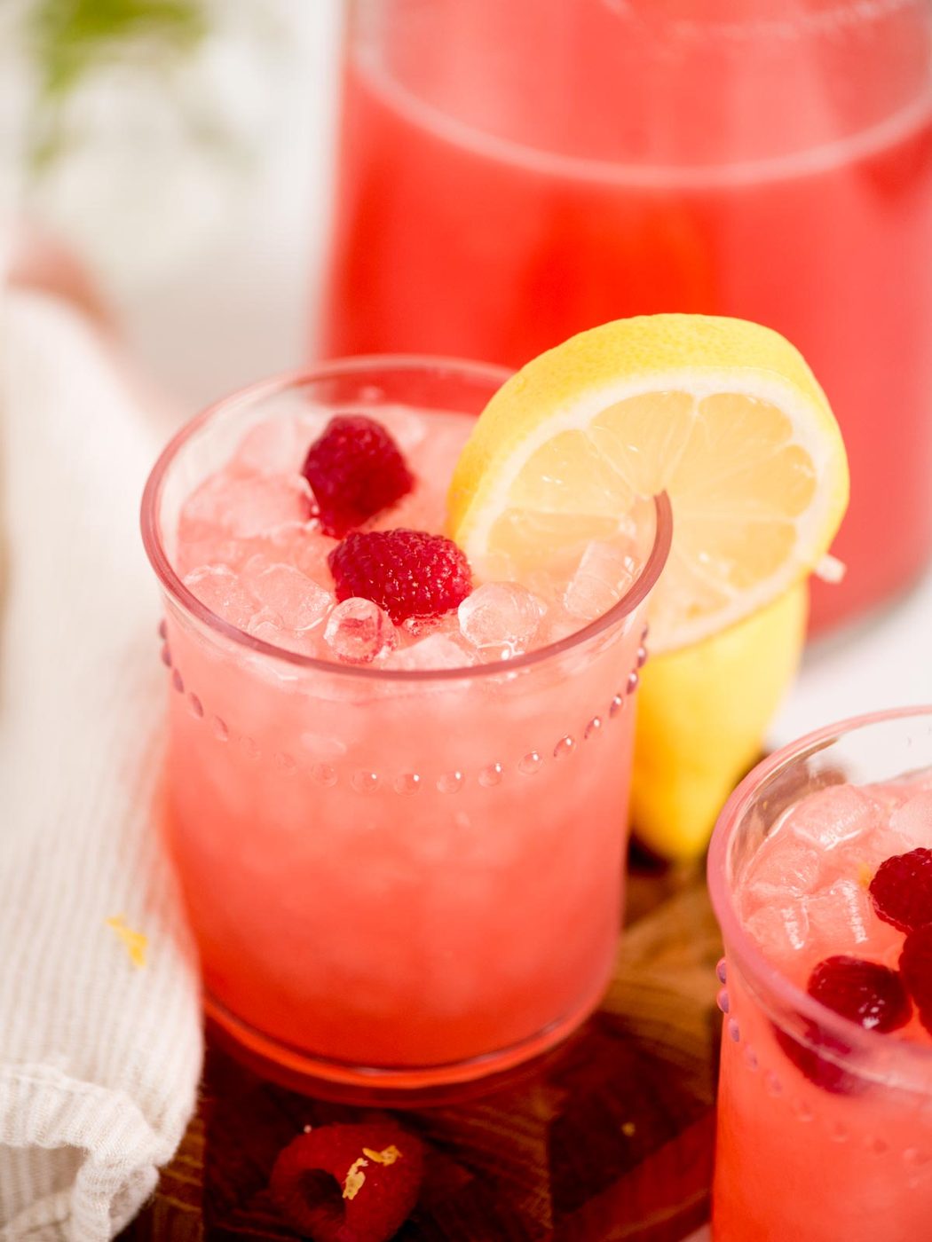 A glass of pink raspberry lemonade with crushed ice, garnished with two raspberries and a lemon slice. A pitcher of the same drink and a white cloth are in the background.