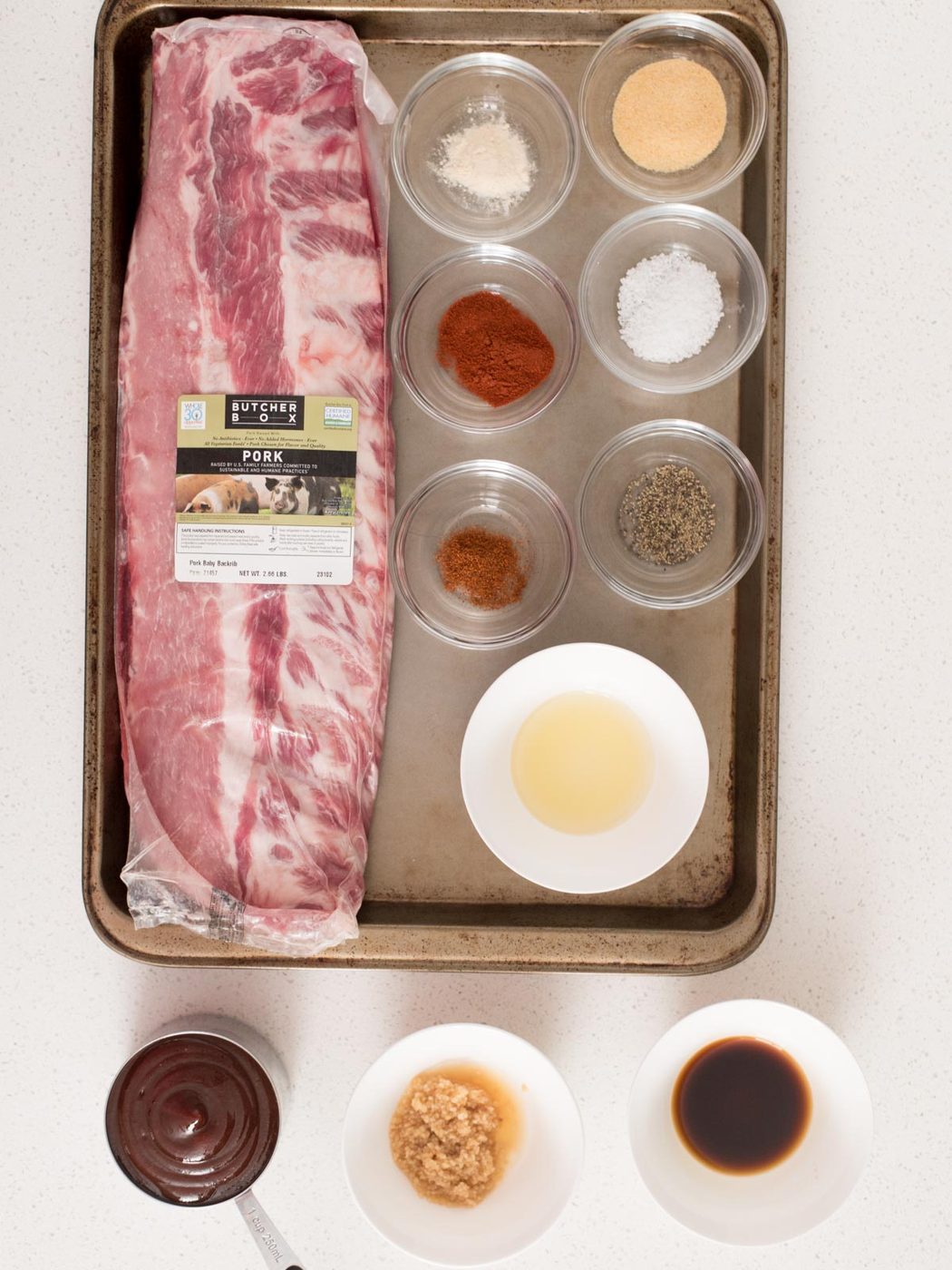 Ingredients used to prepare ribs in the oven measured out in small bowls and displayed on a baking sheet.