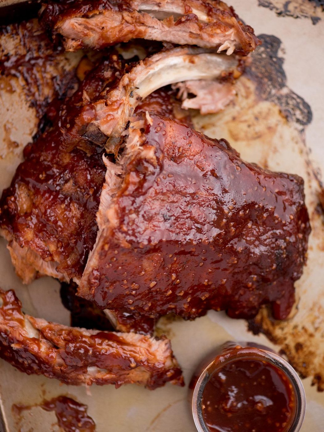 Side of sauce in a bowl on a baking sheet next to tender ribs coated with more sauce, with a few ribs pulled away from the rest.