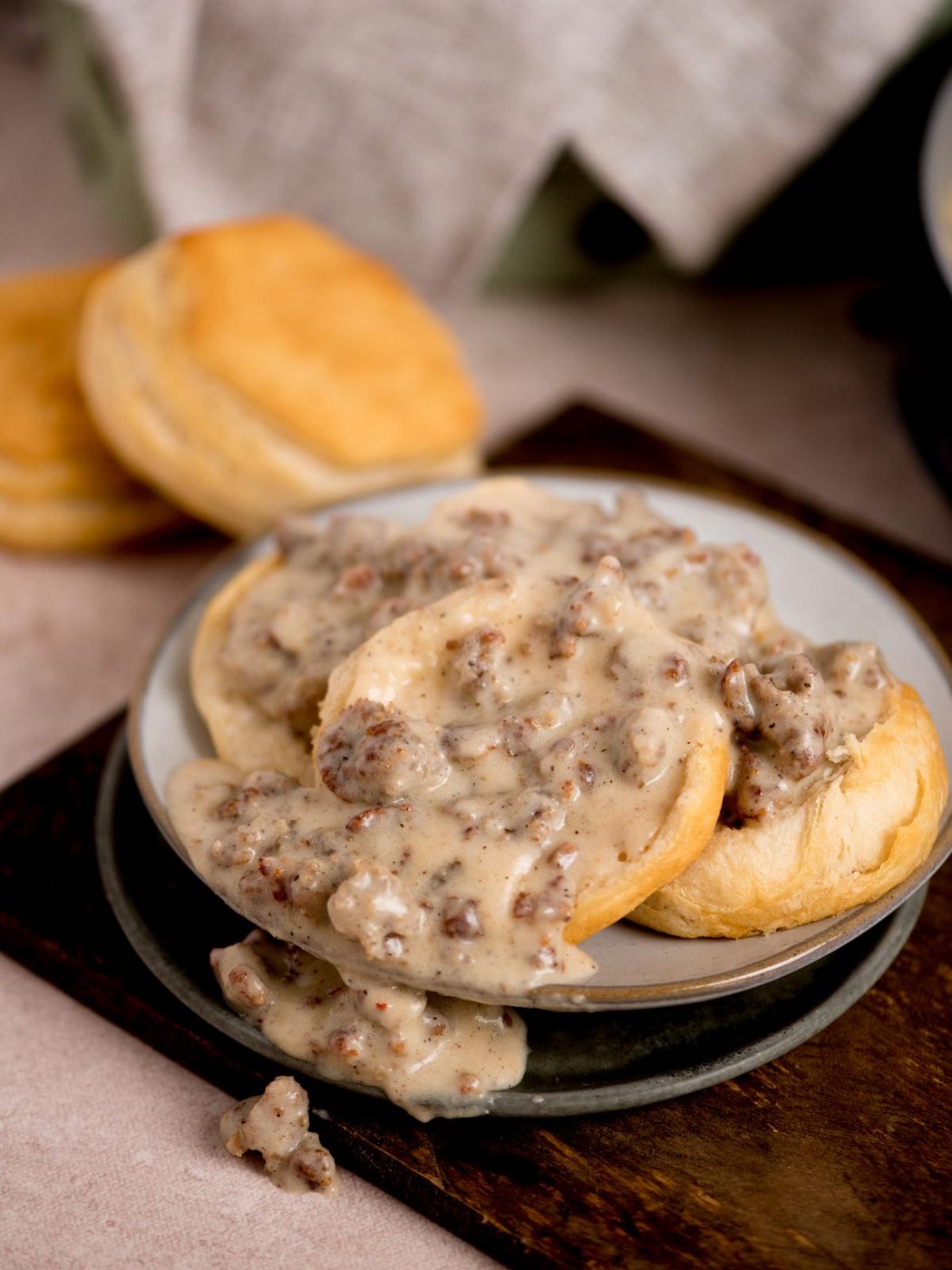 A plate of freshly baked biscuits topped with creamy sausage gravy. Additional biscuits are seen in the background on a wooden surface. The scene suggests a hearty, from-scratch breakfast setting.