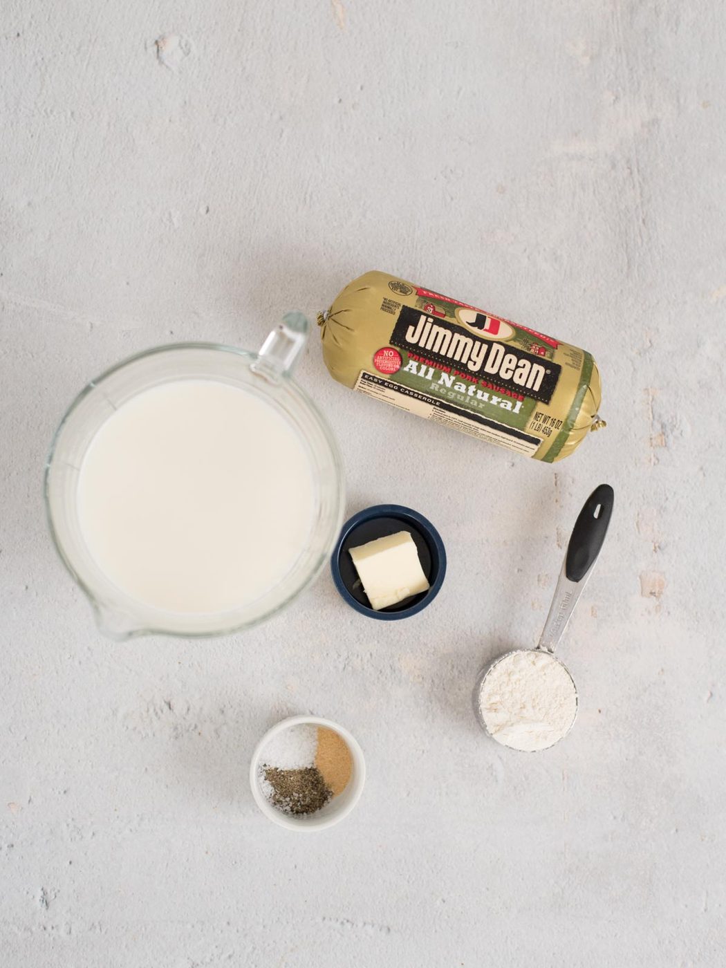 A top view of breakfast gravy ingredients on a light surface: a package of Jimmy Dean sausage, a pitcher of milk, a small bowl with butter, a measuring cup with flour, and a small dish containing salt and pepper.