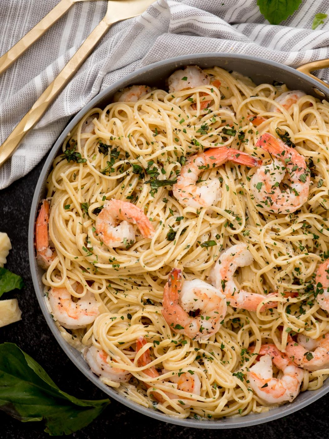 A bowl of shrimp linguine topped with herbs delights the senses. The pasta, luxuriating in a creamy sauce and mingling with succulent shrimp, is garnished with basil leaves and cheese chunks. It rests on a dark surface beside a striped cloth and gleaming golden utensils.