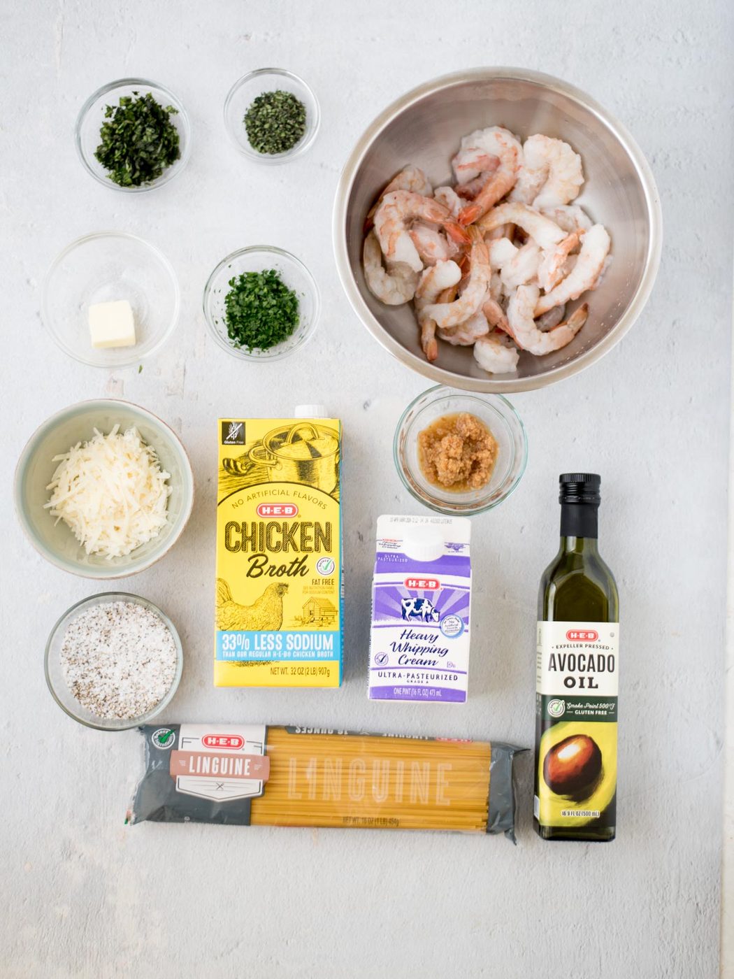 Ingredients for a shrimp linguine dish are artfully arranged on a white surface. Raw shrimp rest in a bowl alongside fresh herbs, grated cheese, and chicken broth. Heavy whipping cream, minced garlic, avocado oil, linguine, and seasoning in small bowls complete the vibrant scene.
