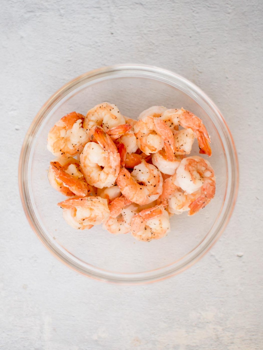 A glass bowl filled with seasoned shrimp, reminiscent of a savory shrimp linguine, sits on a light gray surface.