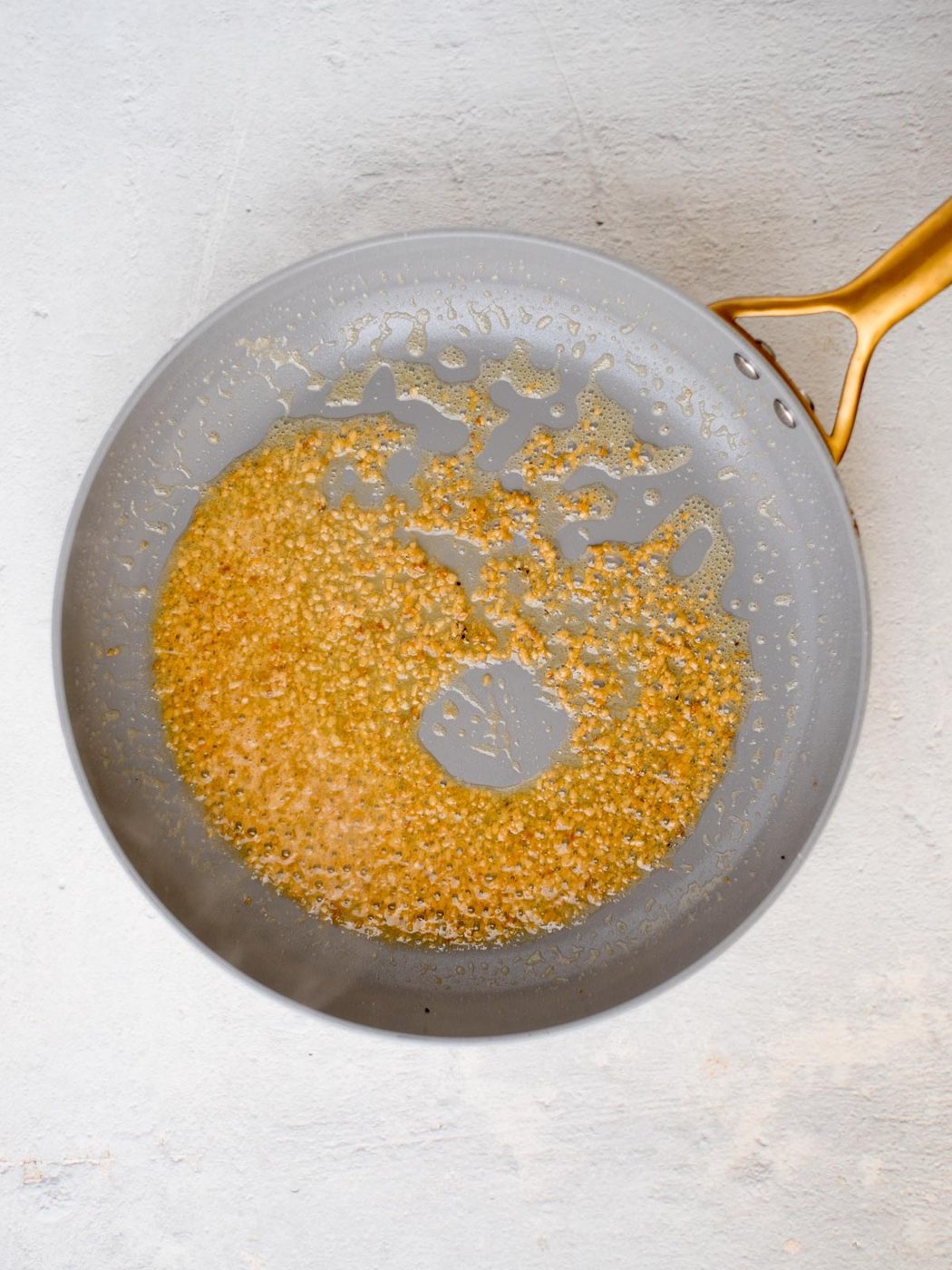A gray frying pan with a gold handle rests on a light surface. Inside, finely minced garlic is being sautéed in oil to a golden-brown hue—evenly prepping the base for a delectable shrimp linguine.