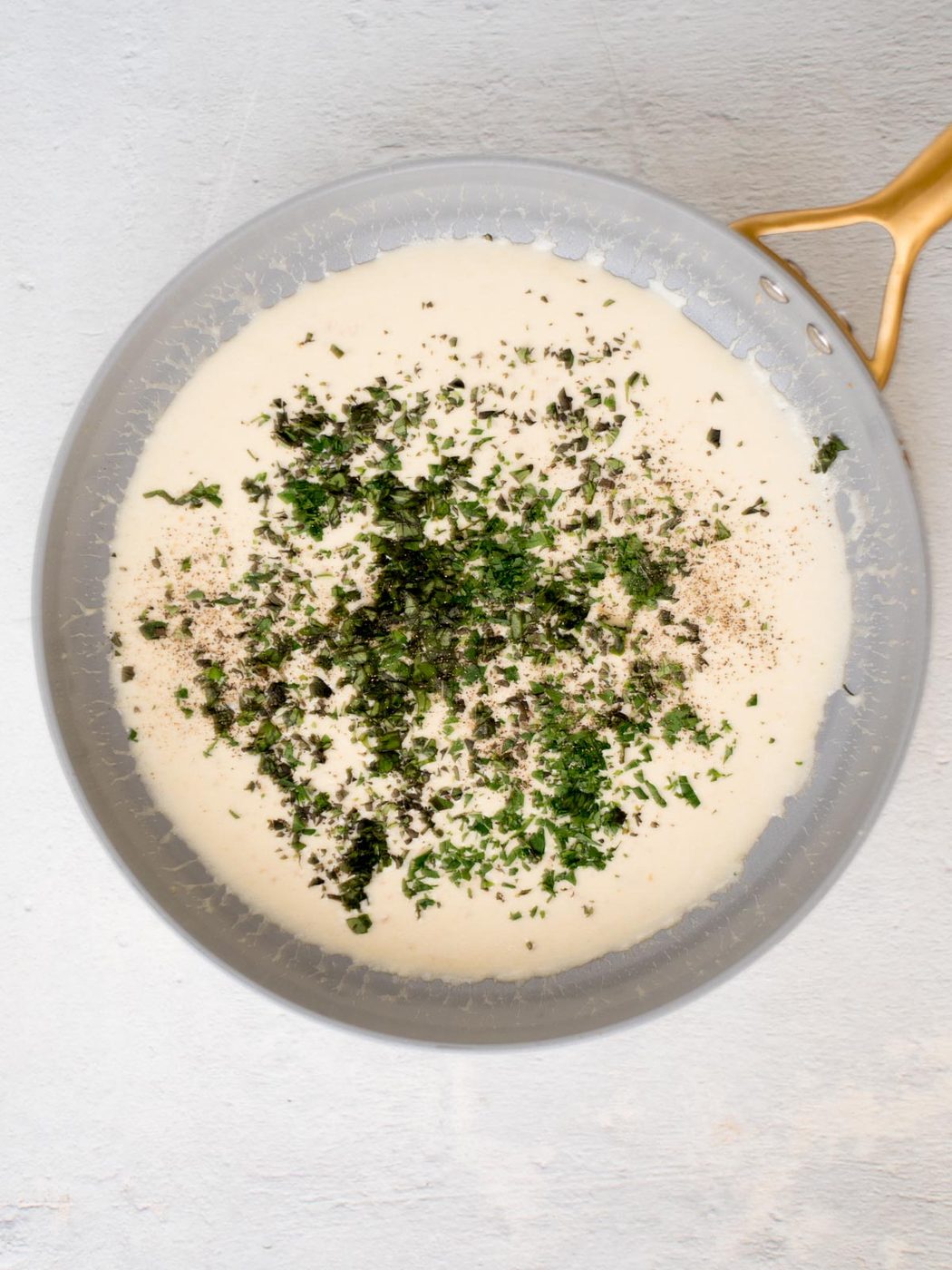 A creamy sauce in a pan with a golden handle, topped with a generous amount of chopped herbs and black pepper. This elegant shrimp linguine features a smooth texture and light color, contrasting beautifully with the green and black specks of seasoning.