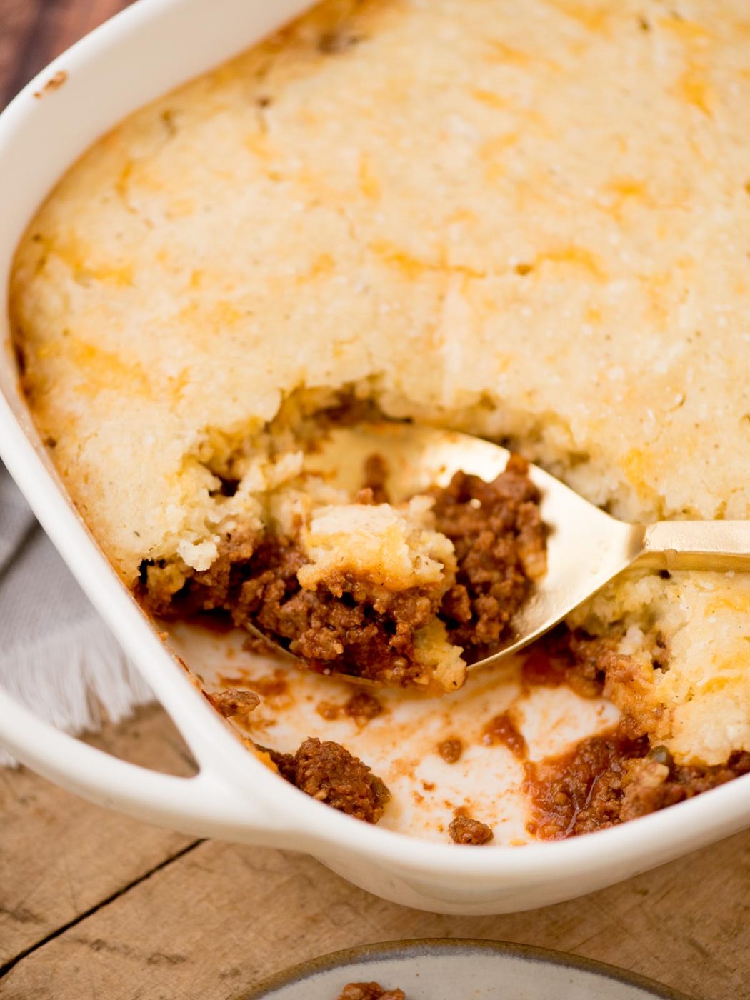 A casserole dish filled with a baked savory pie, featuring a golden crust. A portion has been removed, revealing a layer of minced meat underneath. A serving spoon rests in the dish. The dish is placed on a wooden surface.