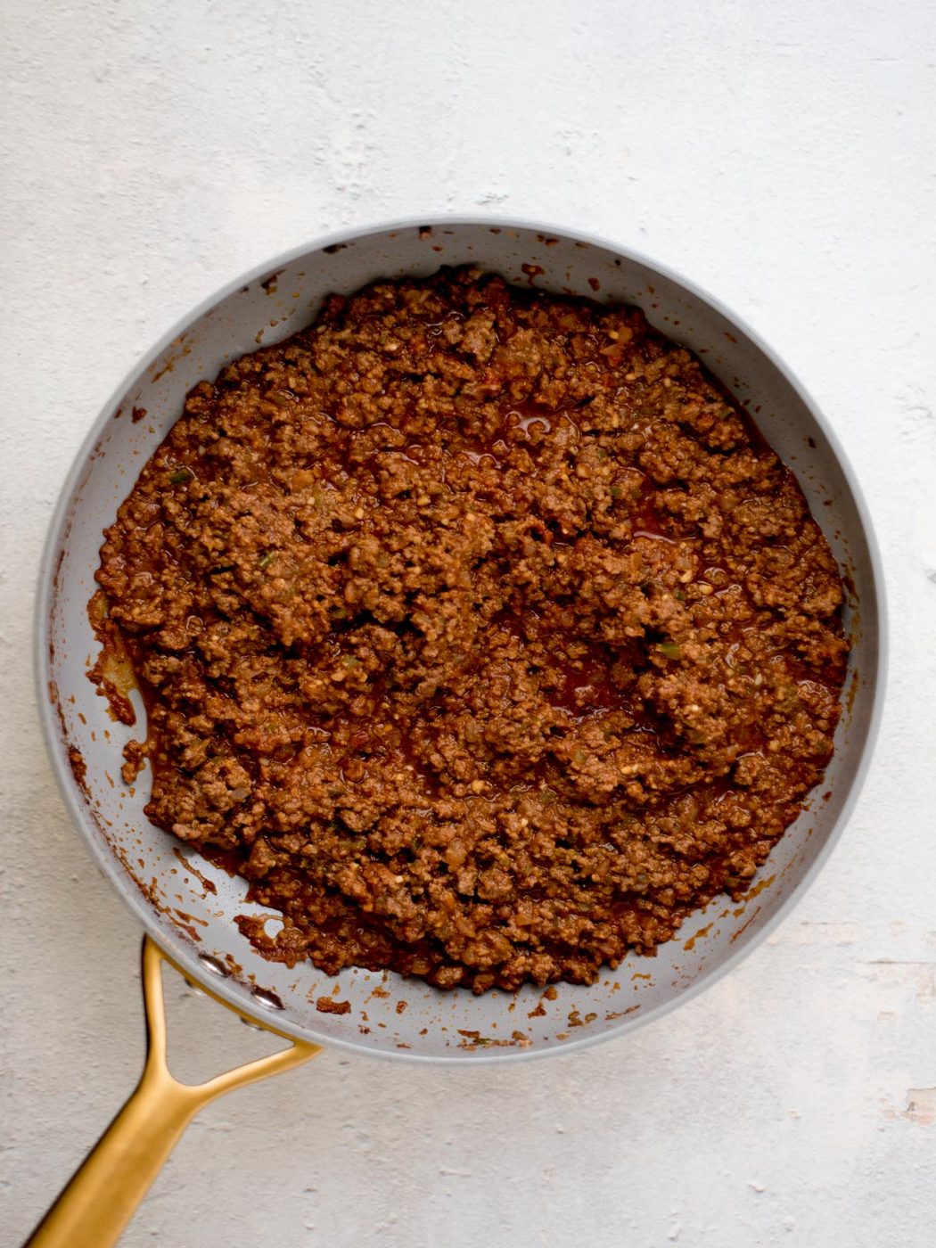 A frying pan filled with seasoned, cooked ground beef, resting on a light-colored surface. The meat is evenly browned, with visible chunks and some sauce mixed in. The pan has a golden handle.