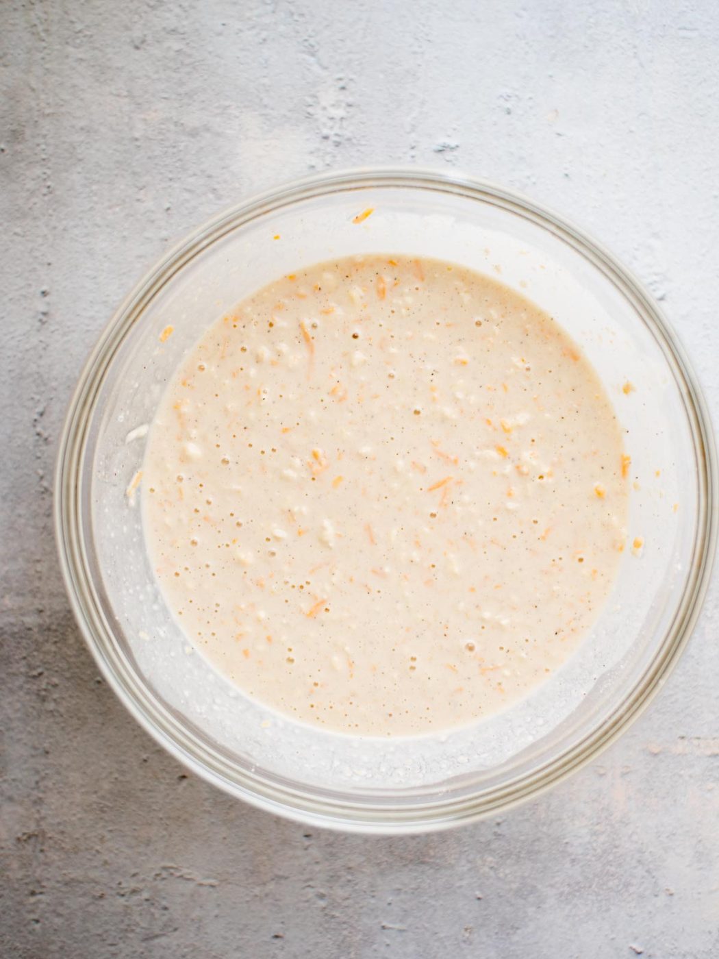 A glass bowl filled with a light beige batter containing visible shreds of orange carrots, sitting on a textured gray surface.