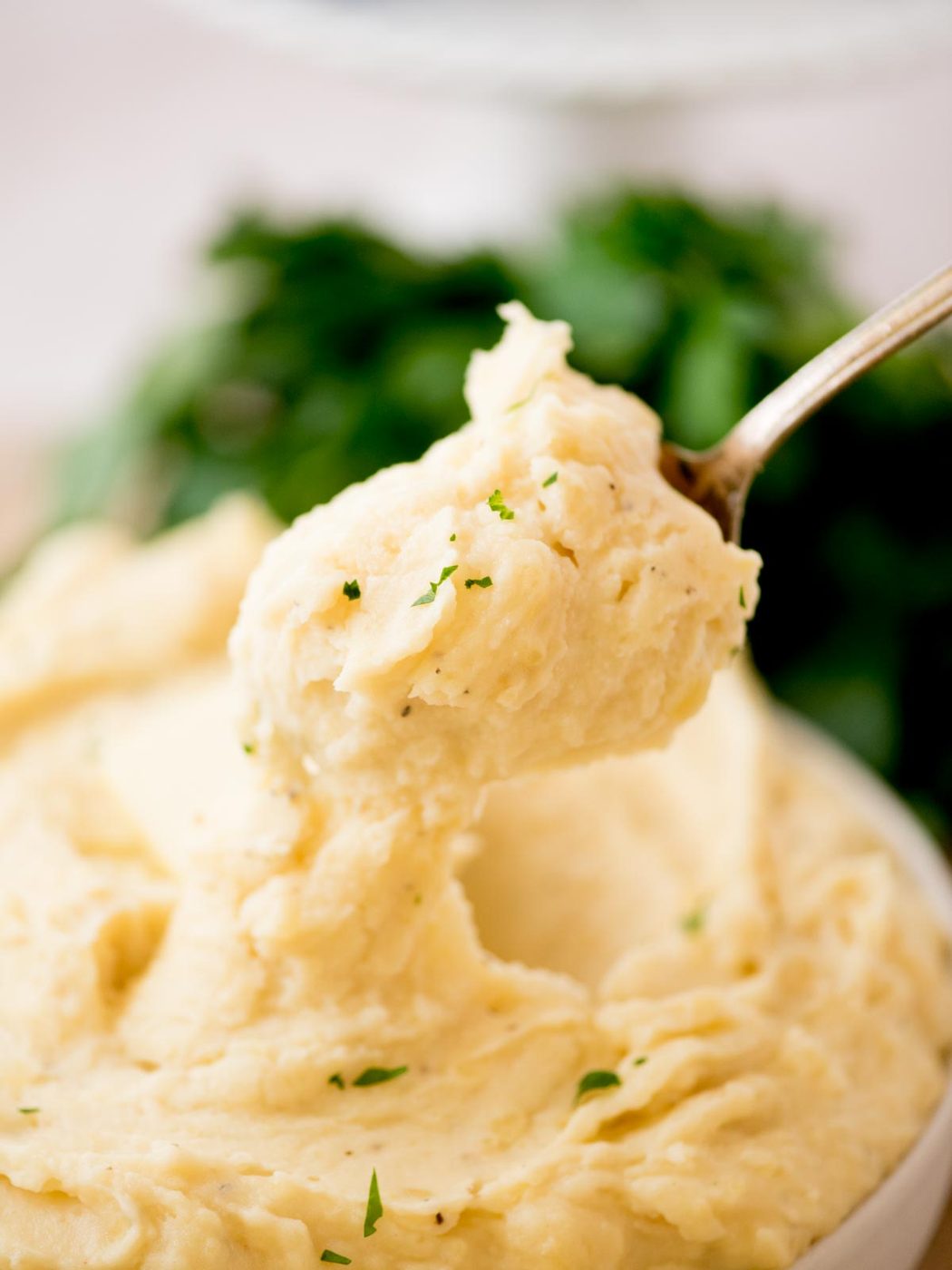 A close-up of creamy slow cooker mashed potatoes being scooped with a spoon, garnished with finely chopped parsley. A blurred green garnish is visible in the background.