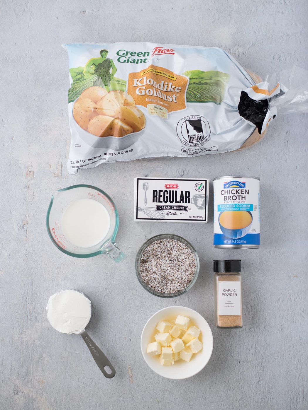 Ingredients for crockpot mashed potatoes laid out on a light surface: a bag of Klondike Goldust potatoes, milk in a measuring cup, cream cheese, chicken broth, a bowl of cubed butter, garlic powder, and a cup of sour cream.