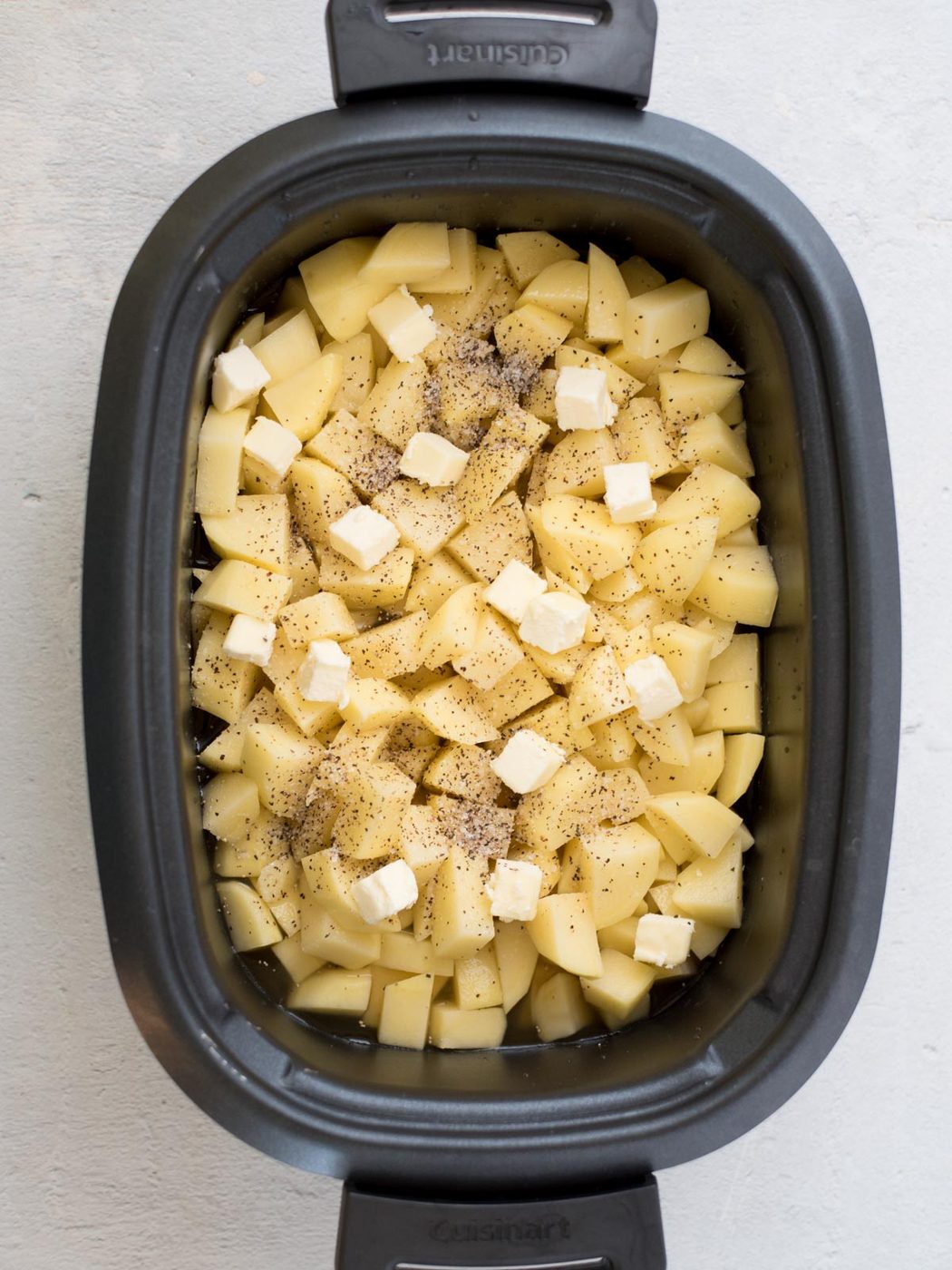 Diced potatoes topped with black pepper and small cubes of butter in a slow cooker, ready to become creamy slow cooker mashed potatoes.