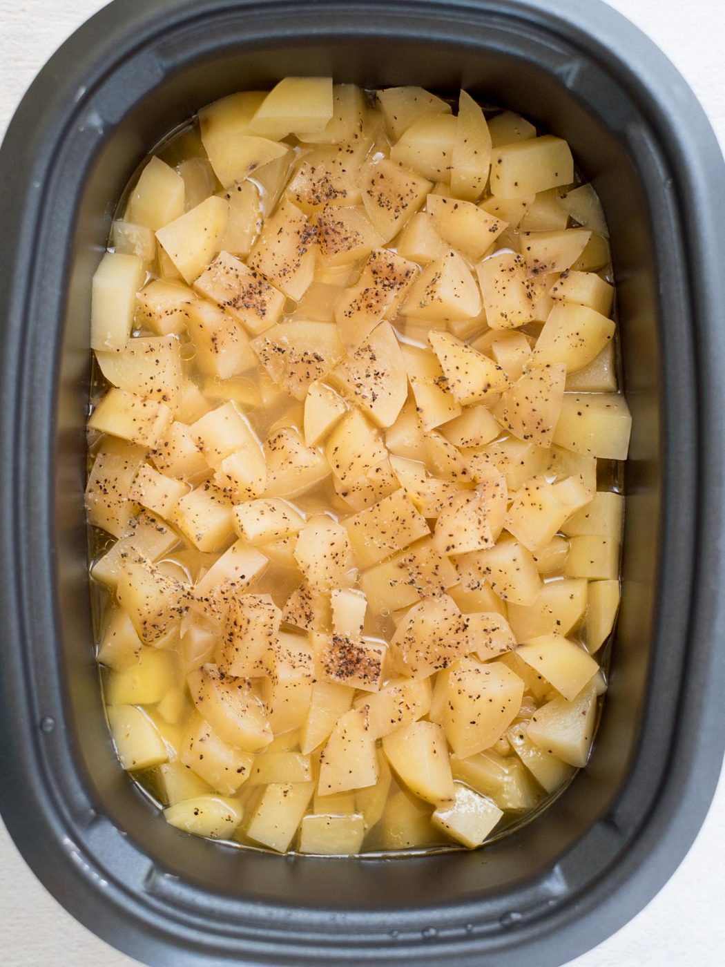 Diced potatoes seasoned with black pepper and cooking liquid inside a slow cooker, suited to making creamy slow cooker mashed potatoes, viewed from above.
