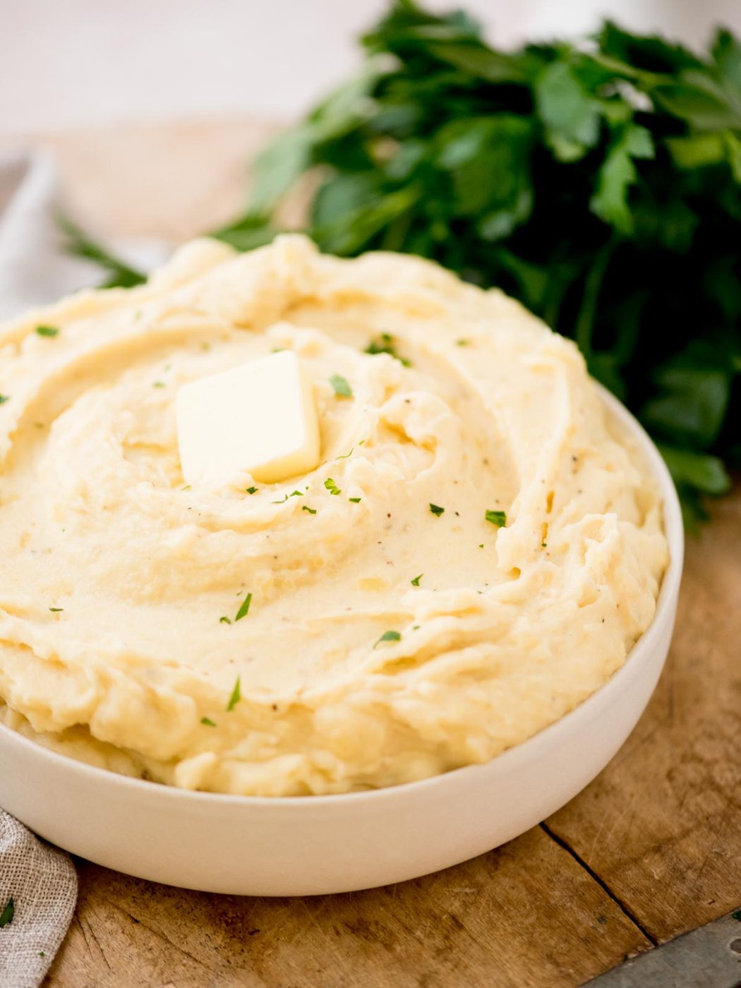 A bowl of creamy slow cooker mashed potatoes topped with a pat of butter and sprinkled with chopped herbs, with fresh parsley in the background.