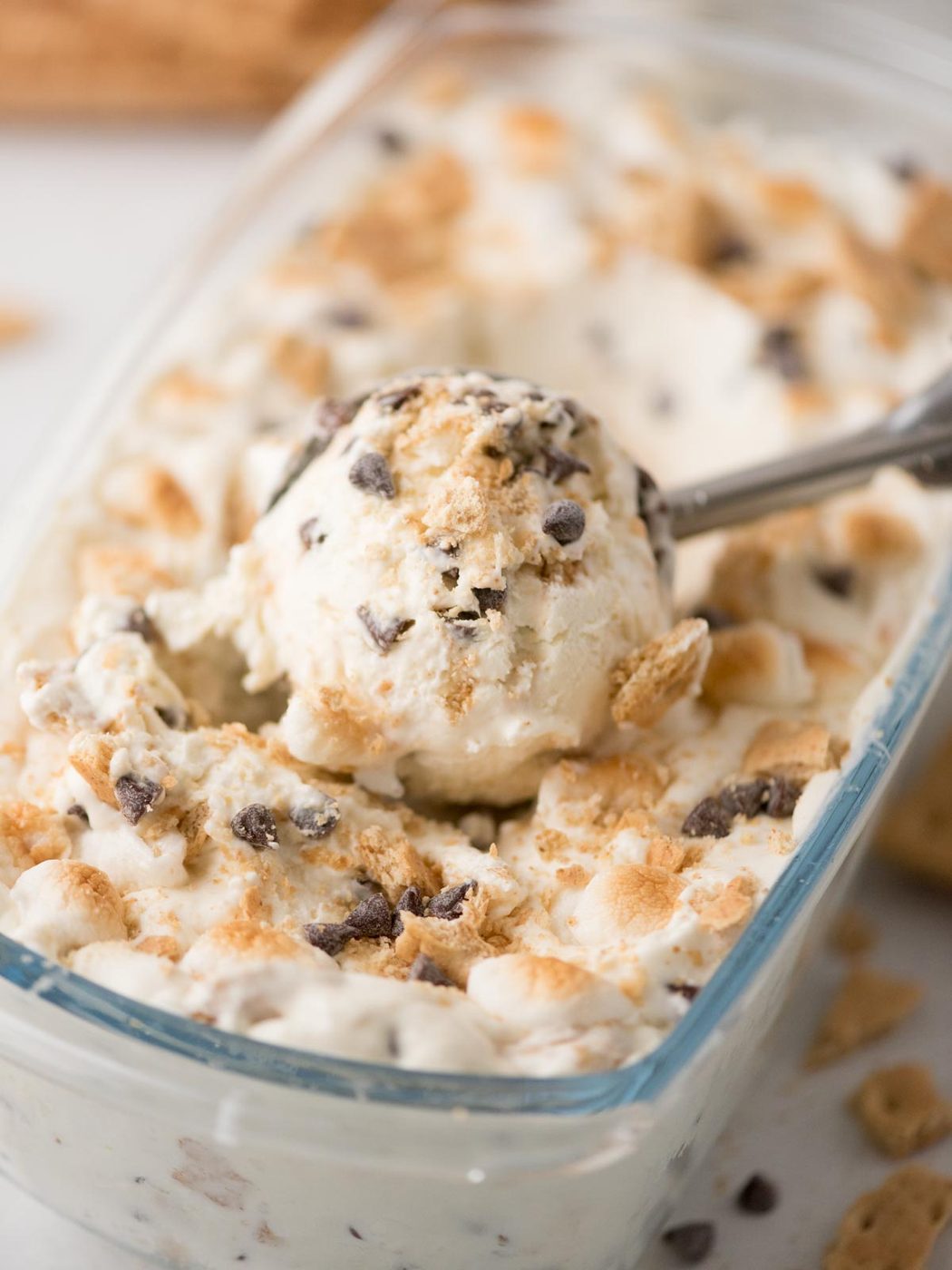 Close-up of a glass dish filled with creamy ice cream mixed with chocolate chips and crushed graham crackers, with a metal scoop lifting a portion.