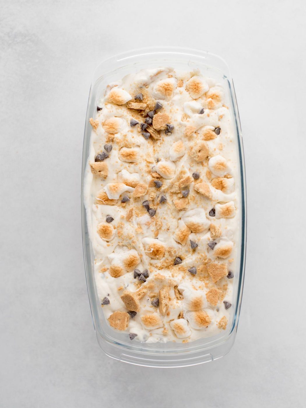 A glass dish filled with ice cream topped with mini marshmallows, chocolate chips, and crushed graham crackers, resembling a smores dessert, on a light gray surface.
